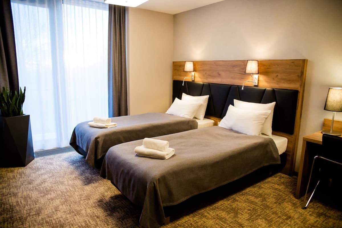 A hotel room with two beds, a large window with curtains, a potted plant, and a desk with a lamp.