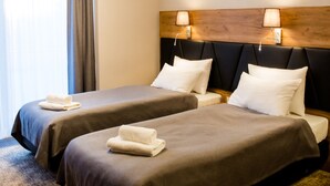 A hotel room with two beds, a large window with curtains, a ceiling air vent, and a headboard with wall-mounted lamps.