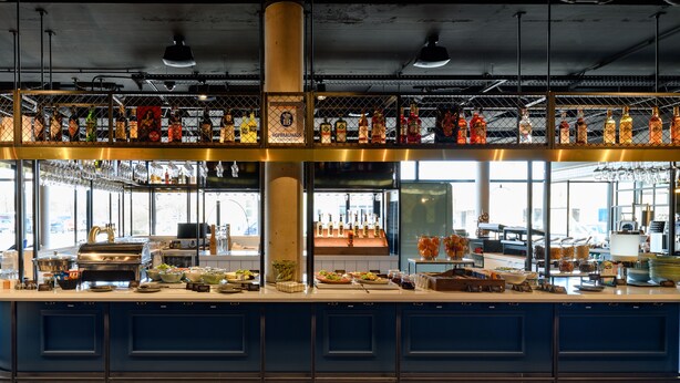 A bar area with a variety of bottles and glasses, a buffet setup with food and drinks, and a patterned floor. - ibis Landshut City (Landshut)