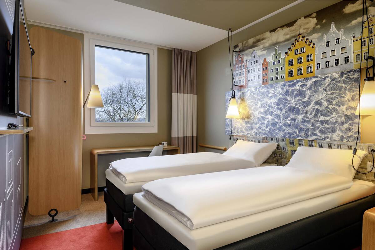 A hotel room with two beds, a window, a stone wall mural, and a TV.