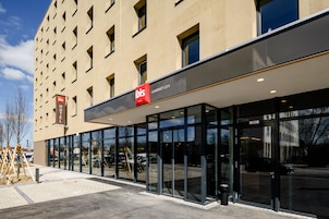 Un hotel moderno con fachada de vidrio, un llamativo letrero rojo y blanco, y vista despejada a la calle.