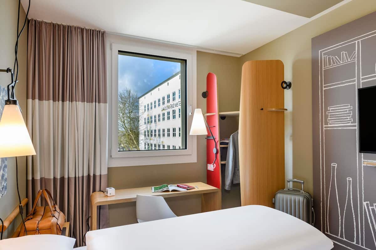 A hotel room with a bed, desk, chair, lamp, window, and a view of a building outside.