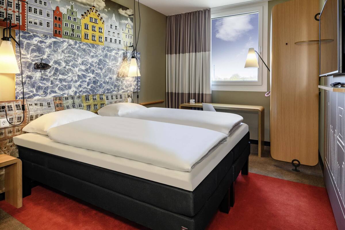 A hotel room with two beds, a mural of buildings, a window with curtains, a desk, and a TV.