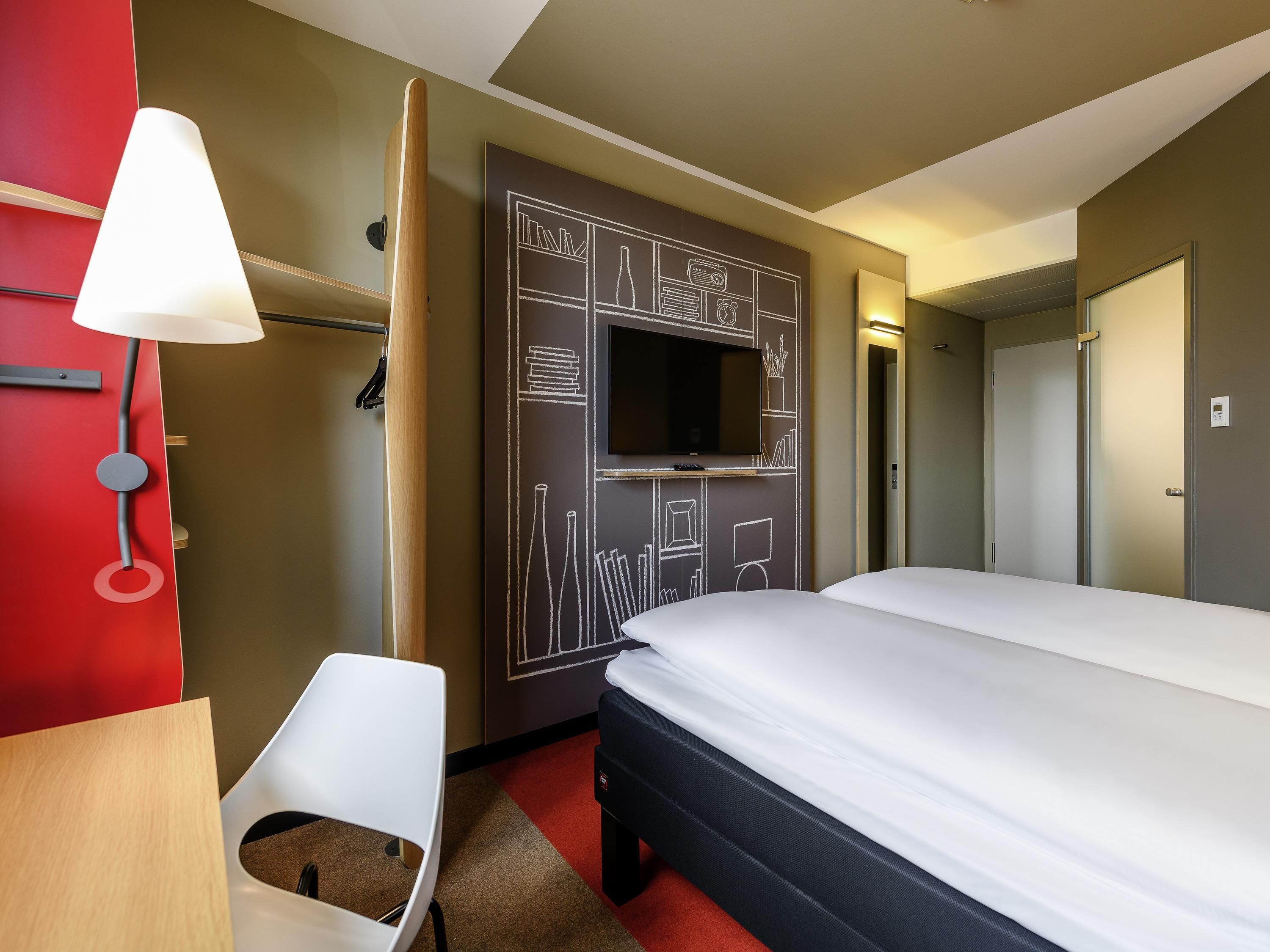 Et hotelværelse med en seng, et tv ophængt på væggen, et skrivebord med en lampe og en stol.