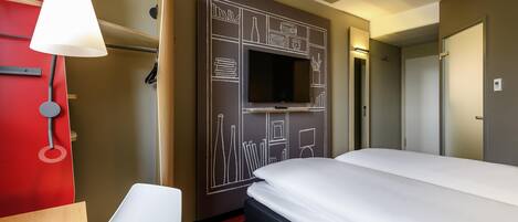A hotel room with a bed, a TV mounted on the wall, a desk with a lamp, and a chair.
