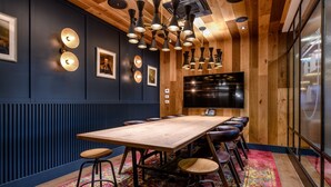 Une salle de conférence avec une longue table en bois, de nombreux tabourets de bar, un tapis coloré et un grand téléviseur à écran plat fixé au mur.