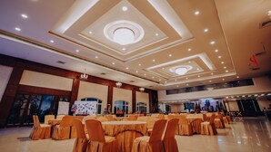 A spacious banquet hall with multiple round tables covered in tablecloths and chairs, a modern ceiling design with recessed lighting, and a large window on the left side.