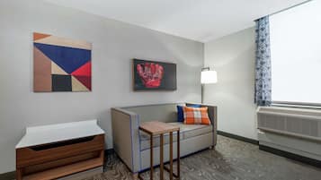 A modern hotel room with a grey sofa, a small wooden table, a wall-mounted artwork, a floor lamp, and a patterned curtain.
