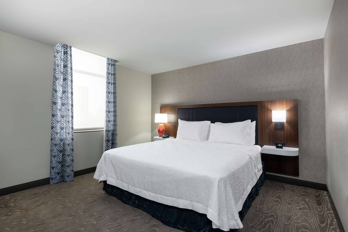 A hotel room with a large bed, two bedside tables, a nightstand, a lamp, and a window with patterned curtains.