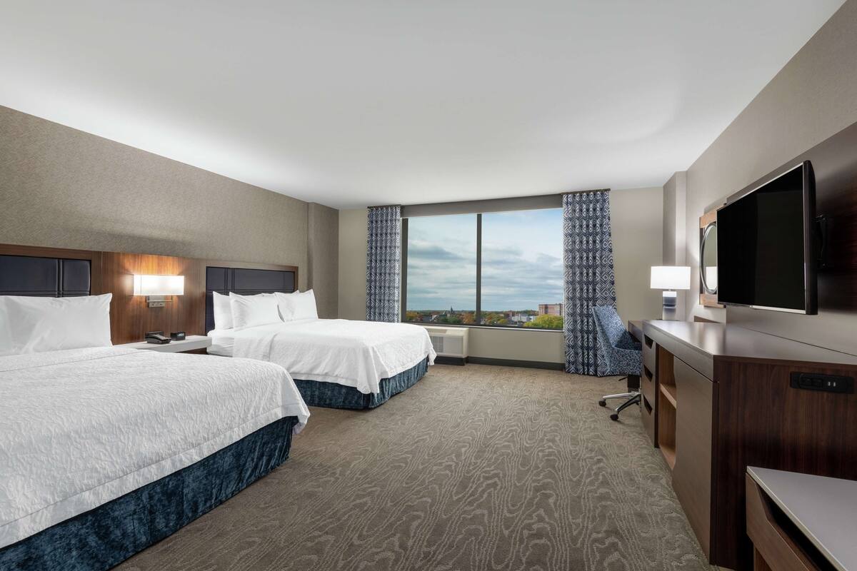 A hotel room with two beds, a desk, a television, and a large window with a view.
