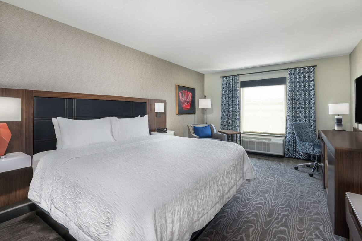 A hotel room with a large bed, a desk, a chair, and a window with patterned curtains.