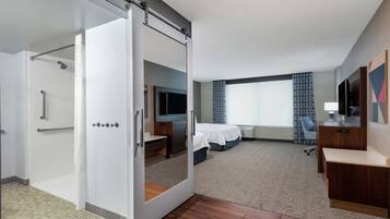 A hotel room with a sliding door to a shower area, a bed with a TV, a desk, and a large window with curtains.
