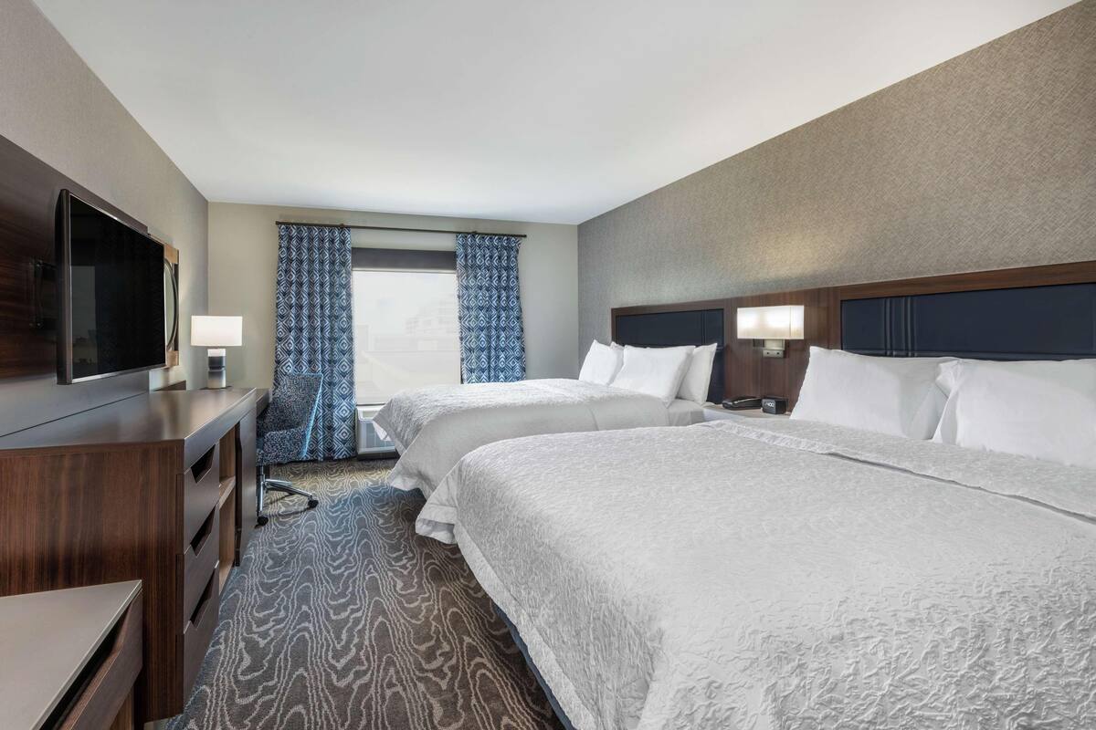 A hotel room with two beds, a TV, a desk, and a window with patterned curtains.