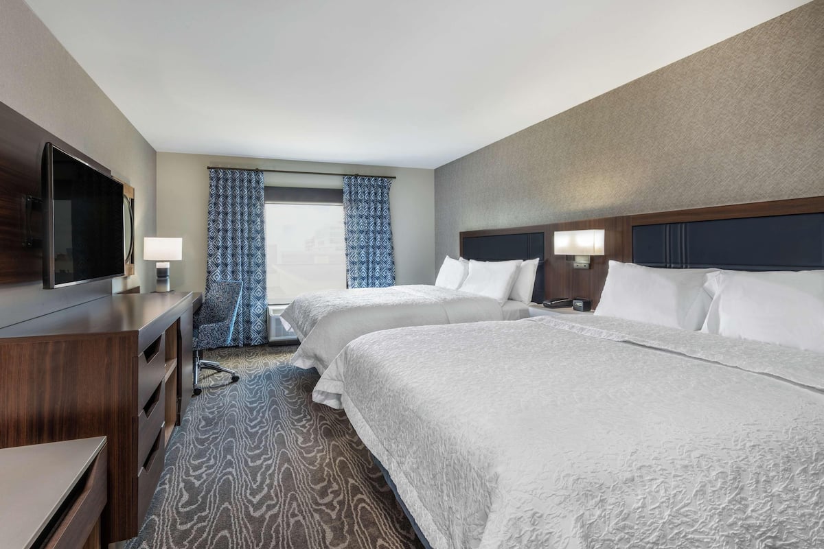 A hotel room with two beds, a TV, a desk, and a window with patterned curtains.
