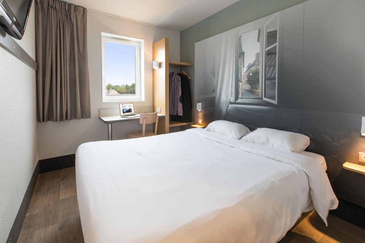 Une chambre d’hôtel avec un grand lit, un bureau avec un ordinateur, une chaise, une armoire et une fenêtre avec des rideaux.