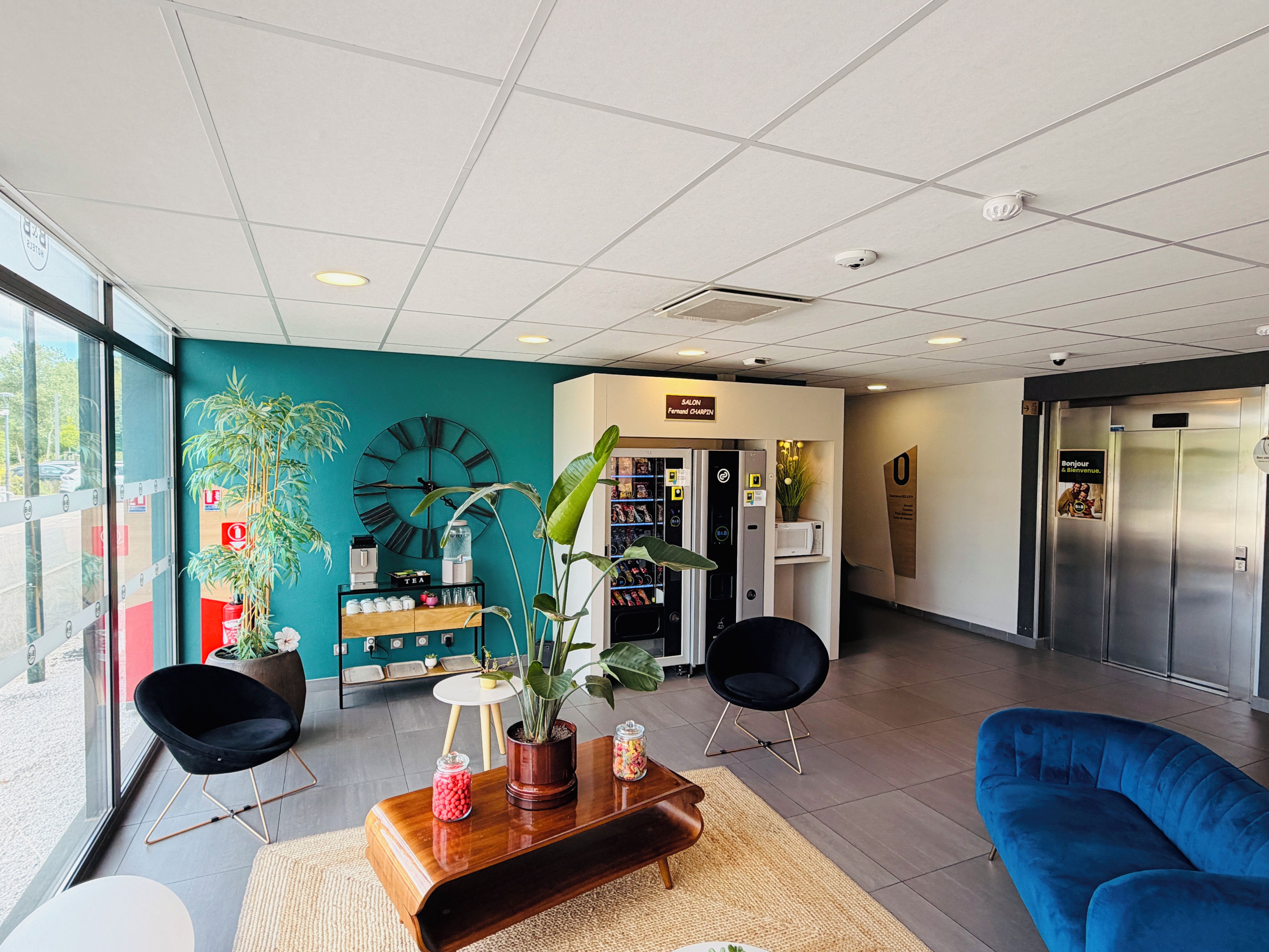 Een moderne lobby met een wand in zachtgroen, een grote klok, een koffiehoek, een receptiebalie en een blauwe bank.