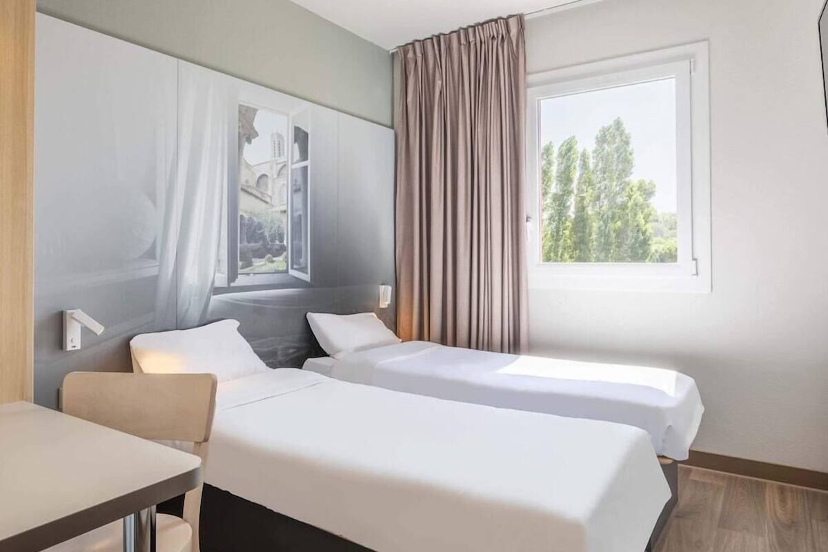 Une chambre d’hôtel avec deux lits, un bureau, une fenêtre avec des rideaux et un luminaire fixé au mur.