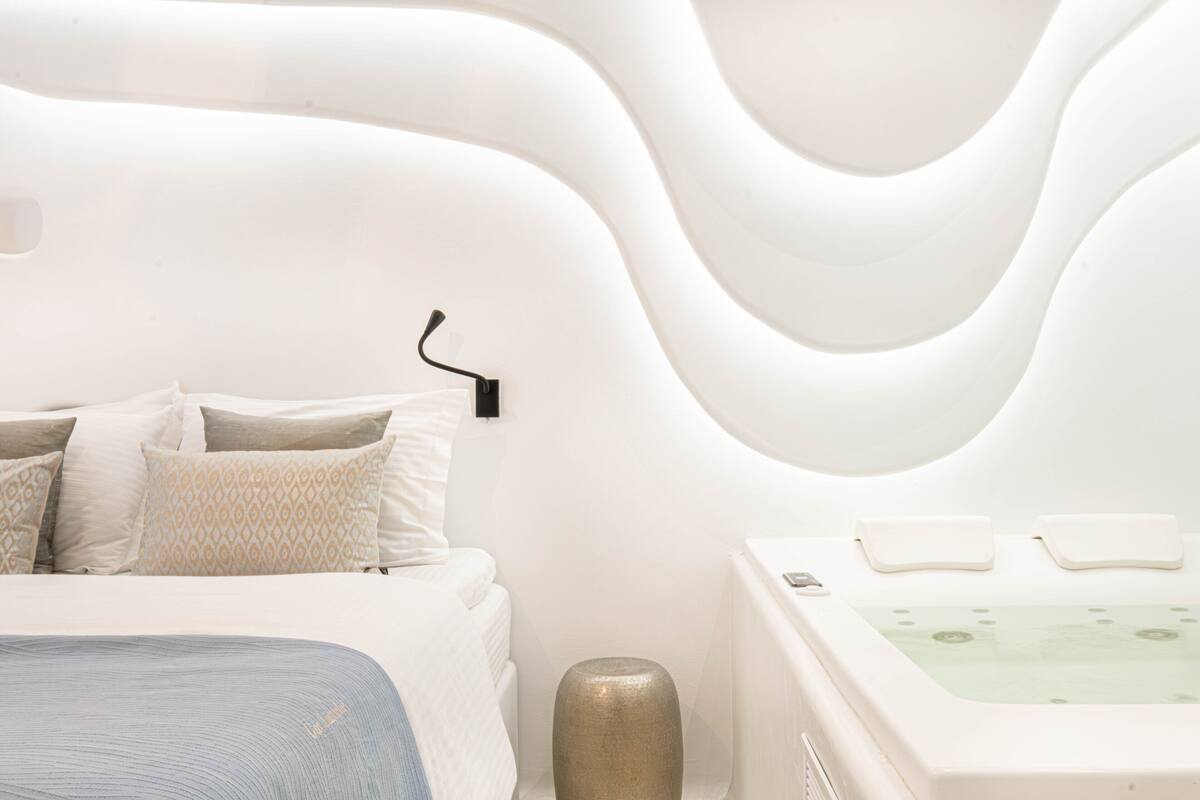 Ein modernes Schlafzimmer mit gewölbter Wandgestaltung, einem Bett mit gemusterten Kissen und einer freistehenden Badewanne.
