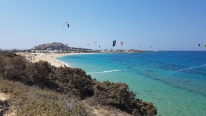 A coastal beach with clear turquoise waters, a sandy shore, and a backdrop of hills with buildings. - AmazeU Naxos (Naxos)
