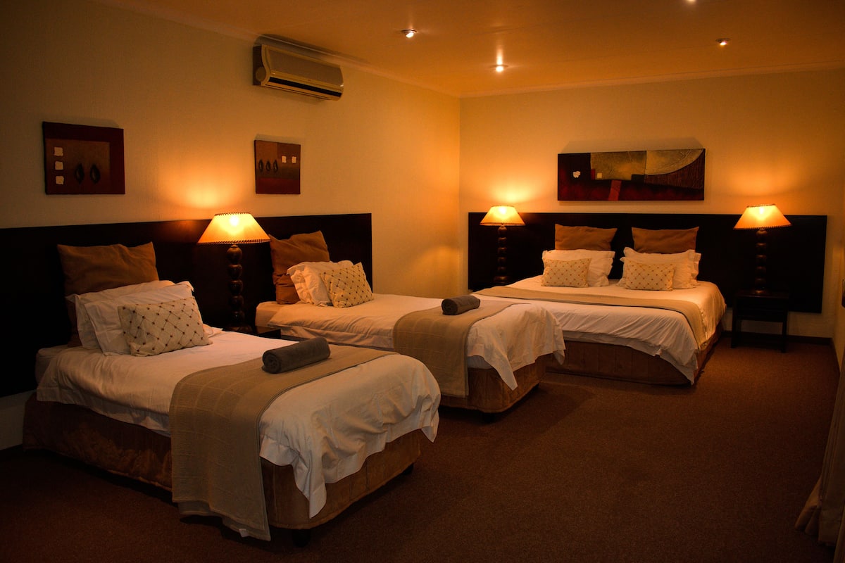 A hotel room with two single beds, each with a nightstand, a lamp, and a headboard. There is an air conditioning unit on the wall and a framed picture above the left bed.