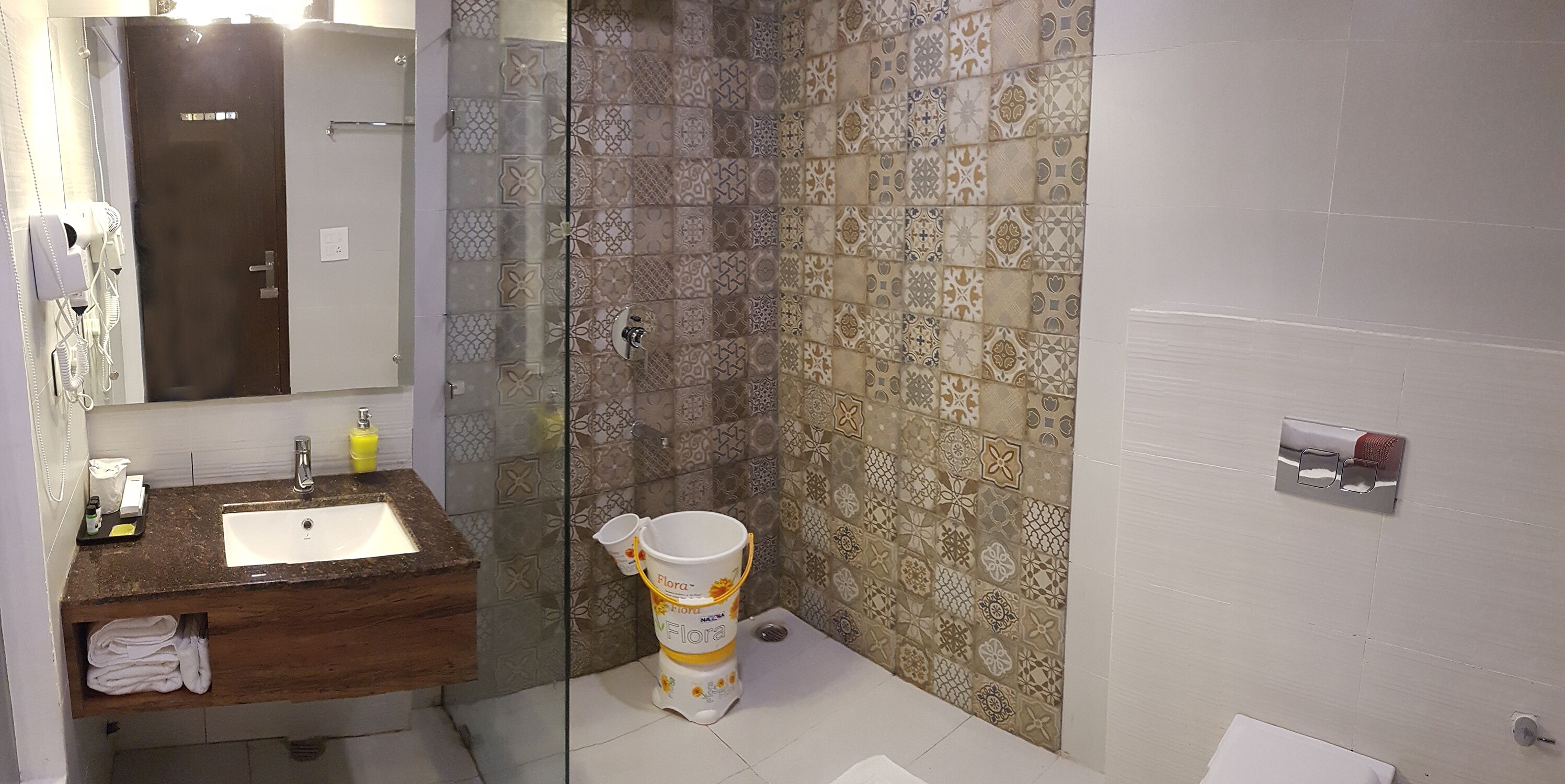 A bathroom with a glass shower enclosure, patterned tiles, a wooden vanity with a sink, and a toilet with a Flora-branded bidet.