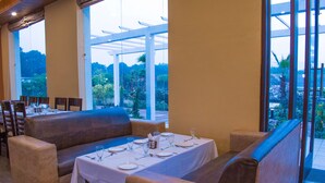 A restaurant with a long table set for dining, cushioned seating, and large windows offering a view of the outdoors. - The Darien Resort (Kaladhungi)