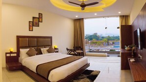 A hotel room with a large bed, a ceiling fan, a TV, and a view of a pool and greenery. - The Darien Resort (Kaladhungi)