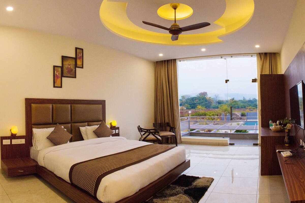 Habitación de hotel con una cama grande, ventilador de techo, televisión y vista a una piscina y vegetación.