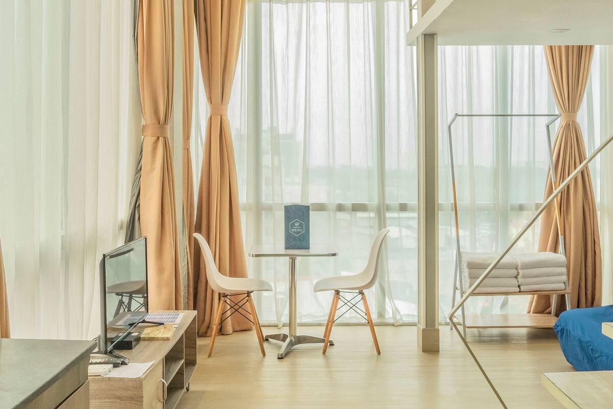A modern hotel room with a desk, two white chairs, a small table with a menu sign, a hanging chair, and large windows with sheer curtains.