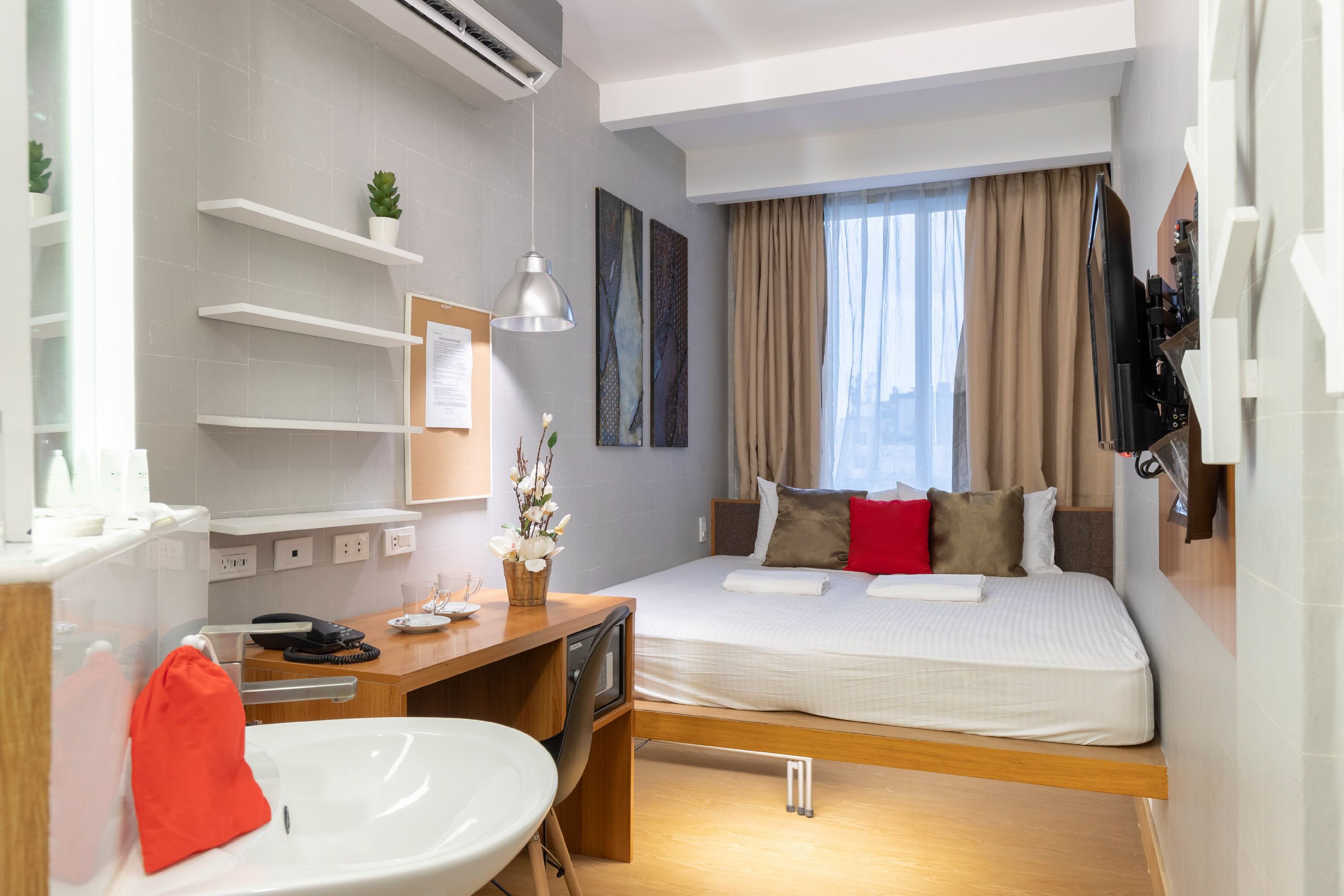A modern hotel room with a wooden desk, a bed with multiple pillows, a wall-mounted shelf with a plant, and a large window with curtains.