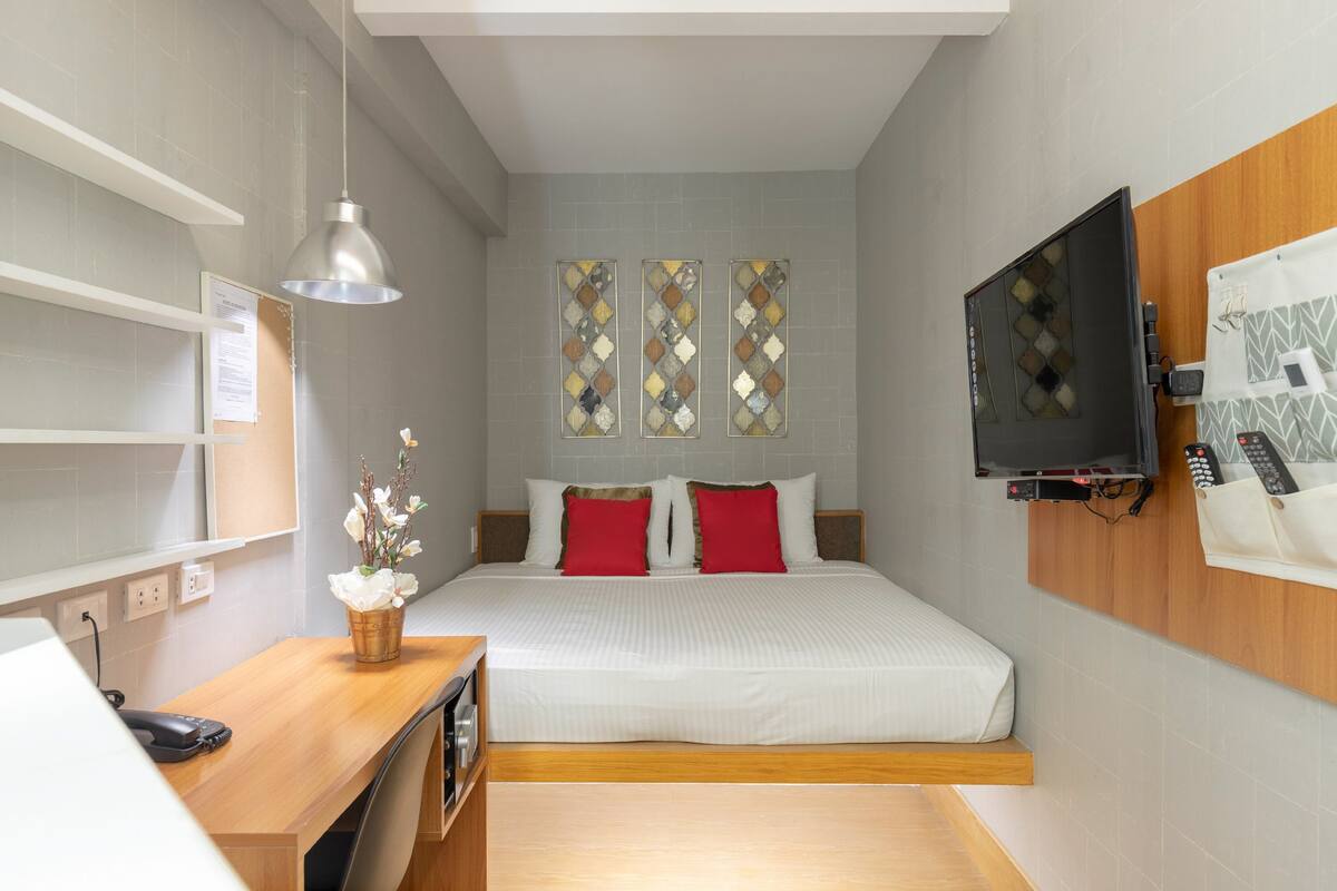 A bedroom with a bed, a desk, a chair, a TV mounted on the wall, and a window with decorative glass panes.
