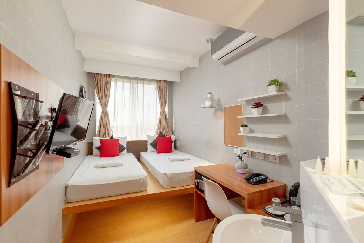 A modern hotel room with two single beds, a desk, a chair, a small table, a mirror, a shelf with plants, and a bathroom with a sink and mirror.