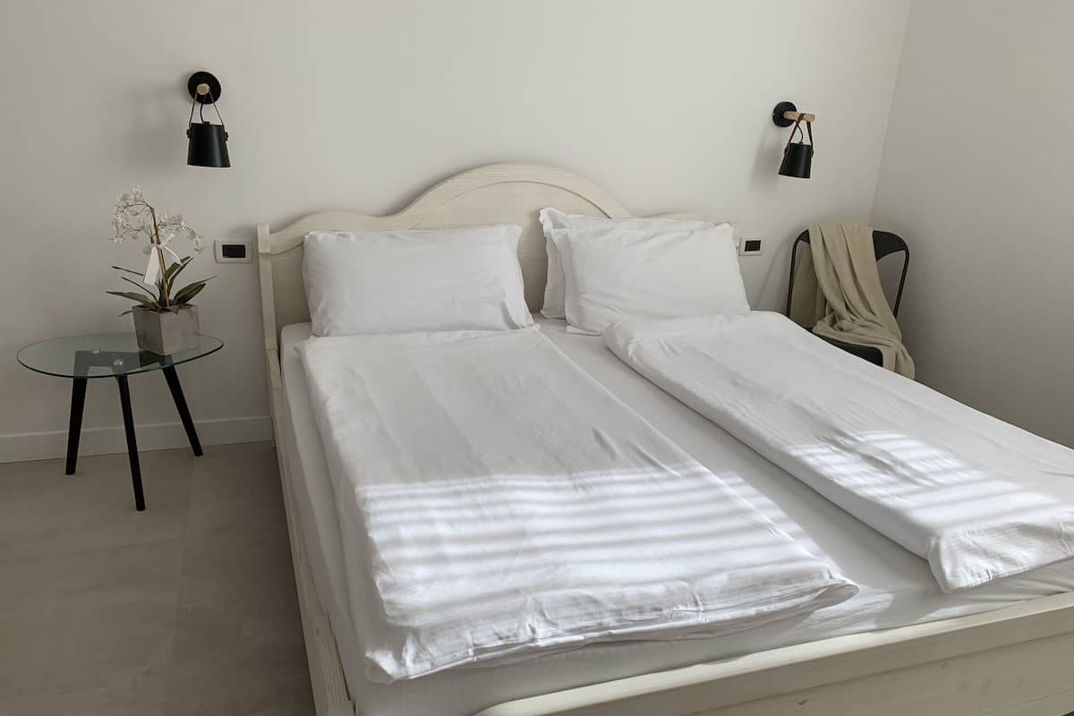 A double bed with white linens, a bedside table with a plant, and two wall-mounted lamps.
