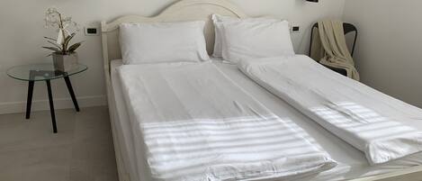 Een tweepersoonsbed met witte lakens, een nachtkastje met een plant, en twee wandlampen.