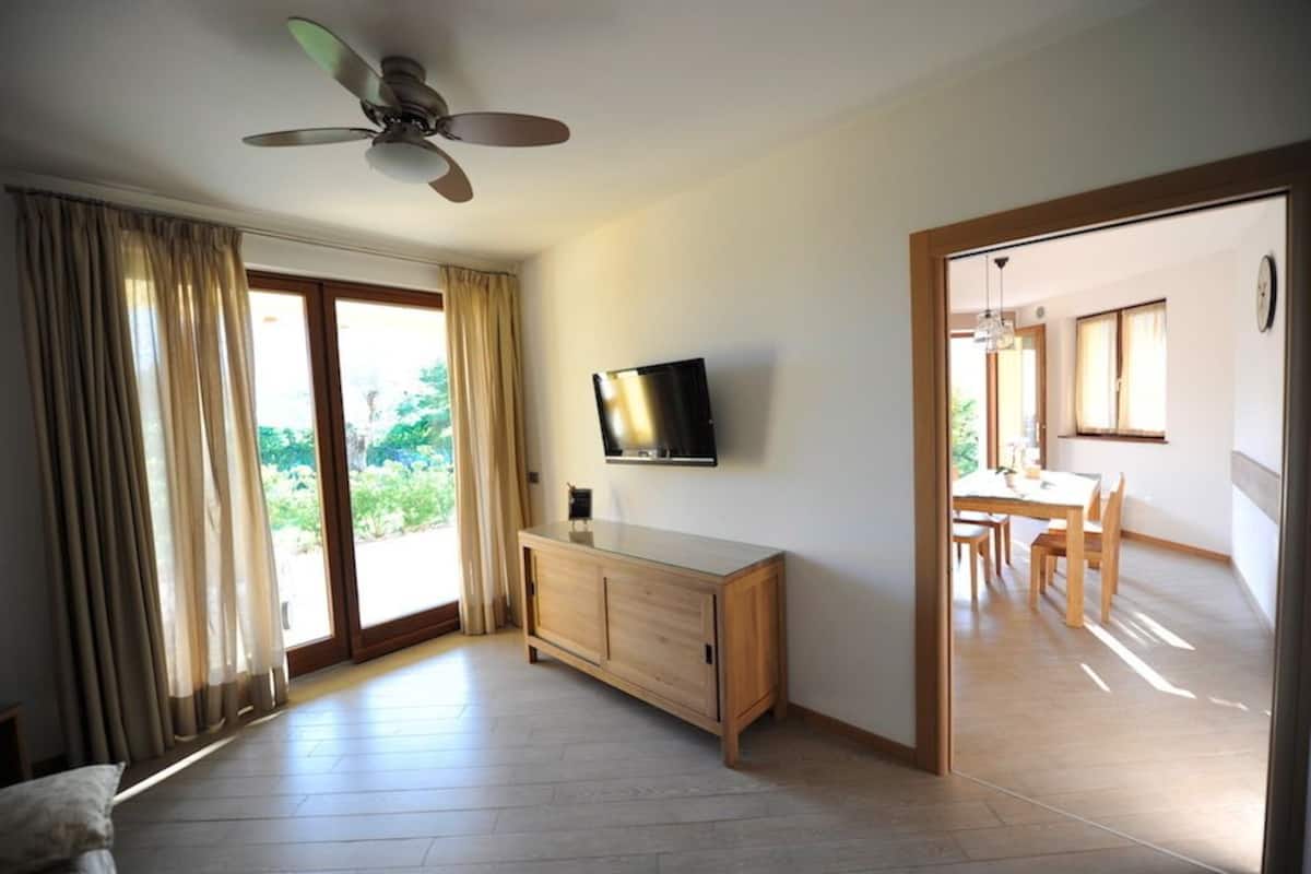 A spacious room with a ceiling fan, a flat-screen TV mounted on the wall, a wooden cabinet, and a sliding glass door leading to a dining area.