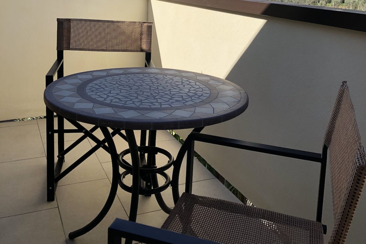 A balcony with a round table and two chairs, overlooking a mountainous landscape with buildings in the distance.