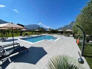 A pool area with lounge chairs and umbrellas, surrounded by a landscaped garden and mountains in the background. - Arcovacanze (Arco)