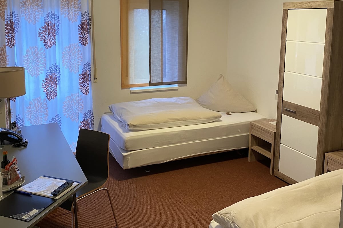 A hotel room with a bed, a desk with a chair, a small table, a window with curtains, and a door.
