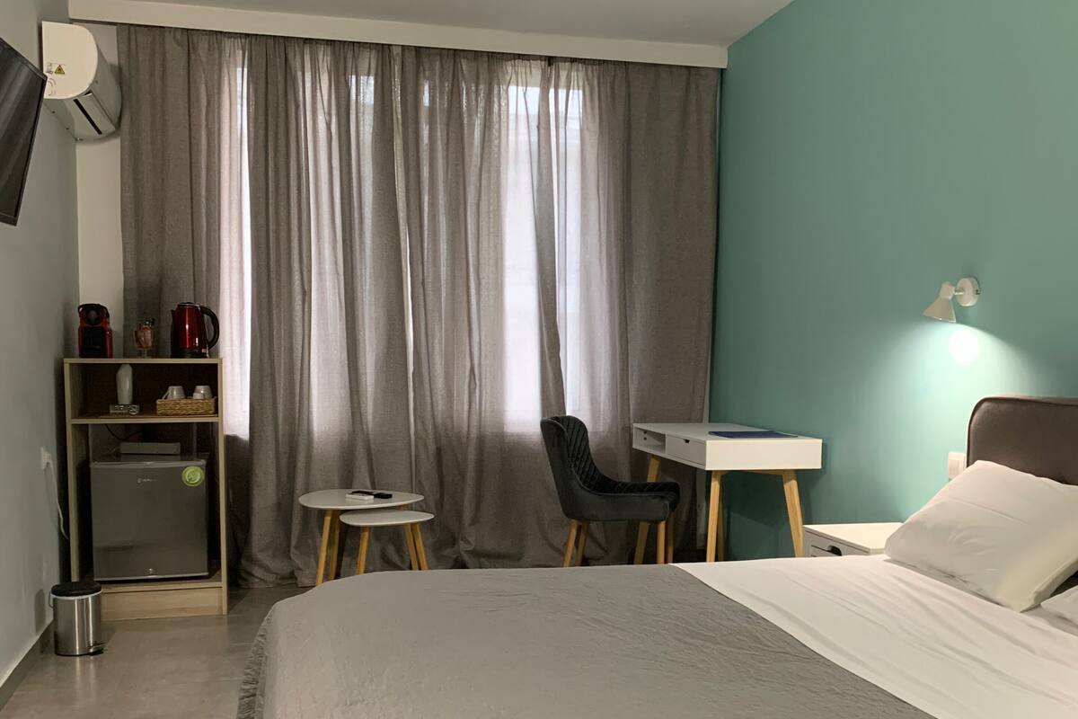 Habitación de hotel con cama, escritorio, silla, mesita, estante con objetos y ventana con cortinas.