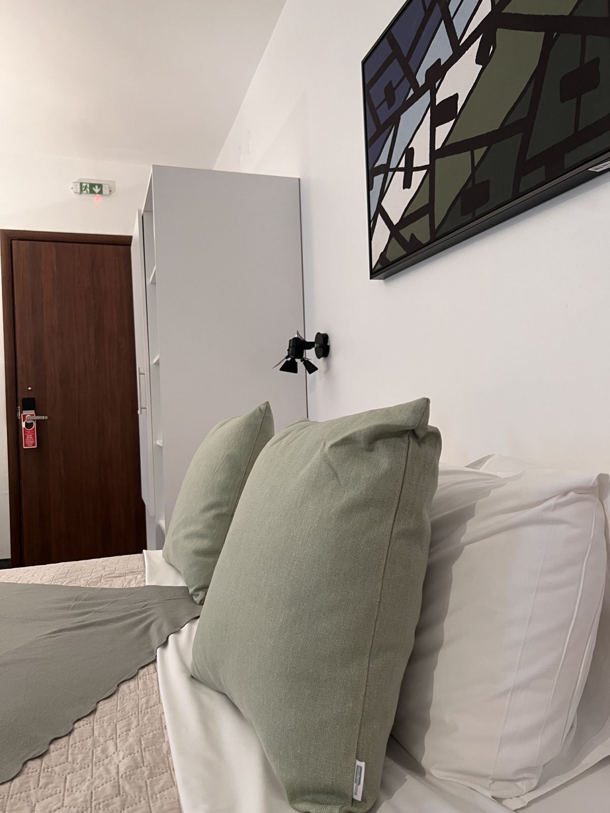 Ein Hotelzimmer mit Bett, Kleiderschrank, Wandleuchte und einem geometrischen Kunstwerk.