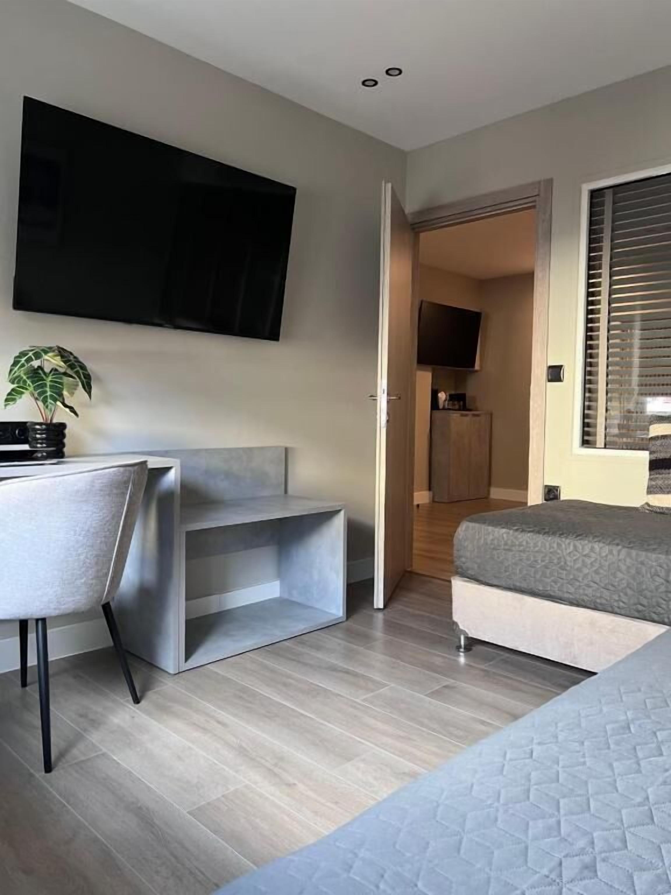 Una habitación moderna con cama, escritorio, silla, televisión montada en la pared y puerta que da a una zona de cocina.