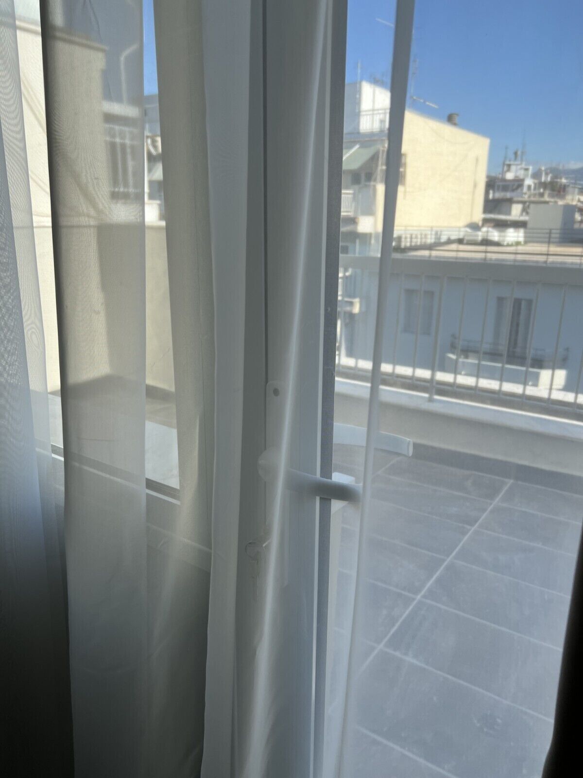Vista a través de una ventana con cortina blanca, mostrando una terraza en la azotea con suelo de baldosas y un edificio al fondo.