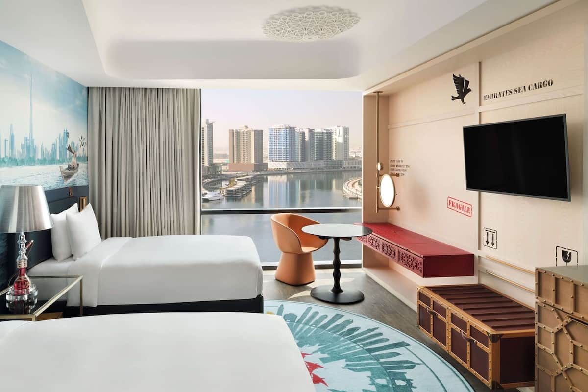 Ein Hotelzimmer mit einem Bett, zwei Nachttischen, einem Fernseher, einem Schreibtisch, einem Sessel und Blick auf die Stadtlandschaft.