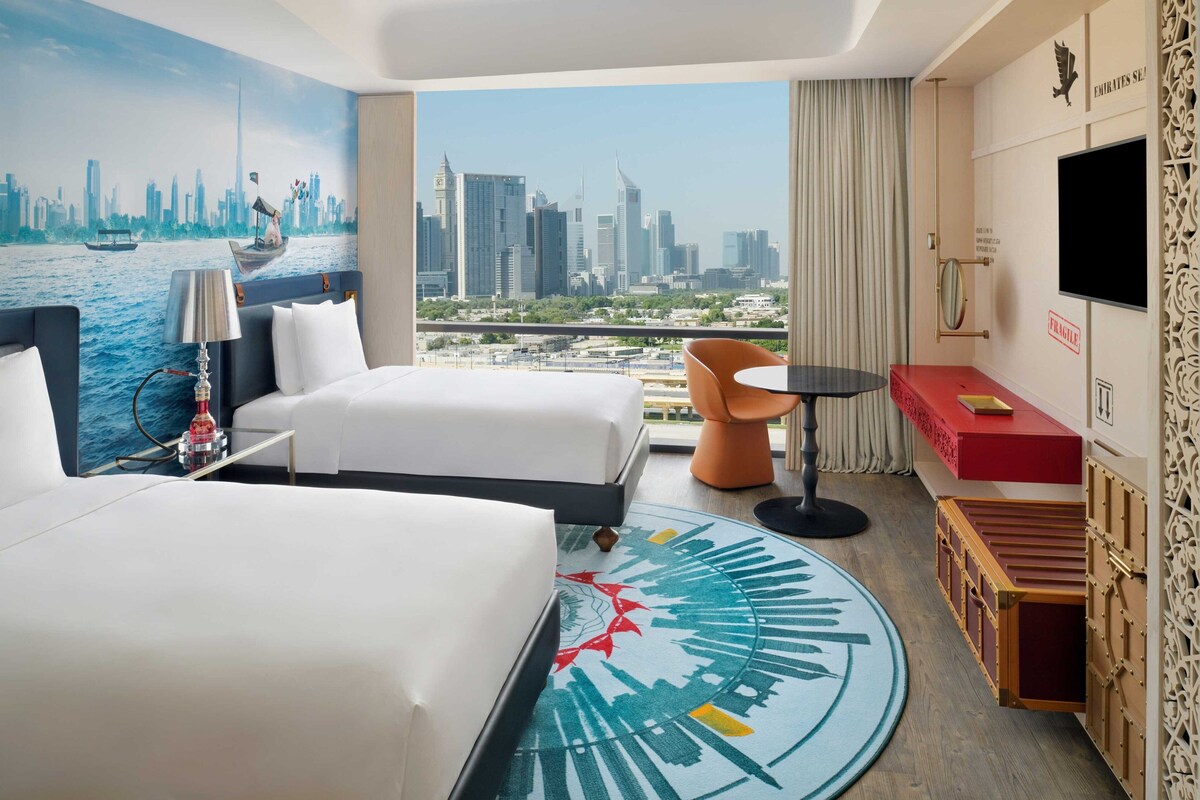 Ein Hotelzimmer mit zwei Betten, einem Stadtbild-Wandgemälde, Blick auf die Stadtlandschaft, einem kleinen Tisch und einem an der Wand befestigten Fernseher.