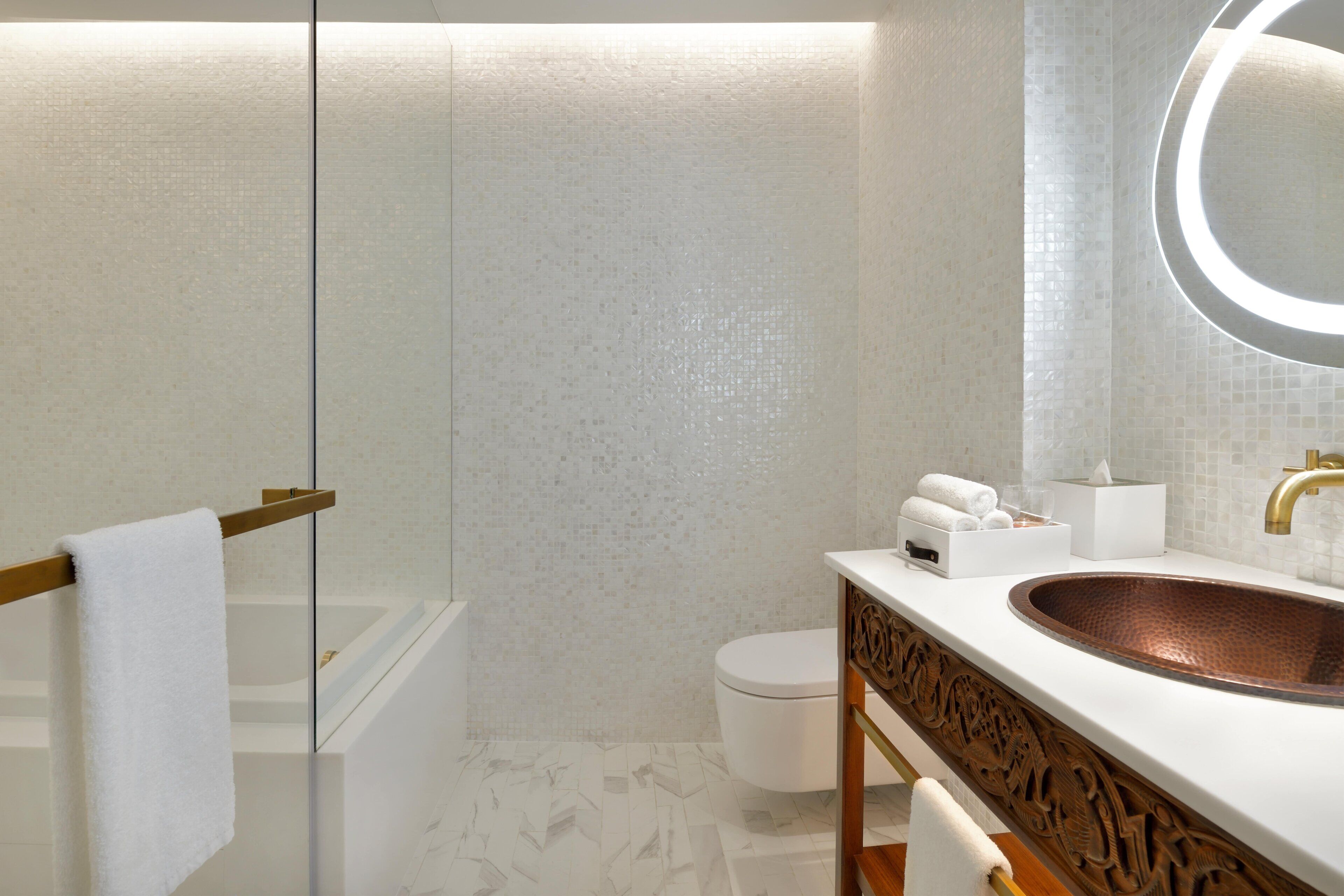 A modern bathroom with a glass shower enclosure, a white toilet, a copper sink, and a mirror with a circular light.