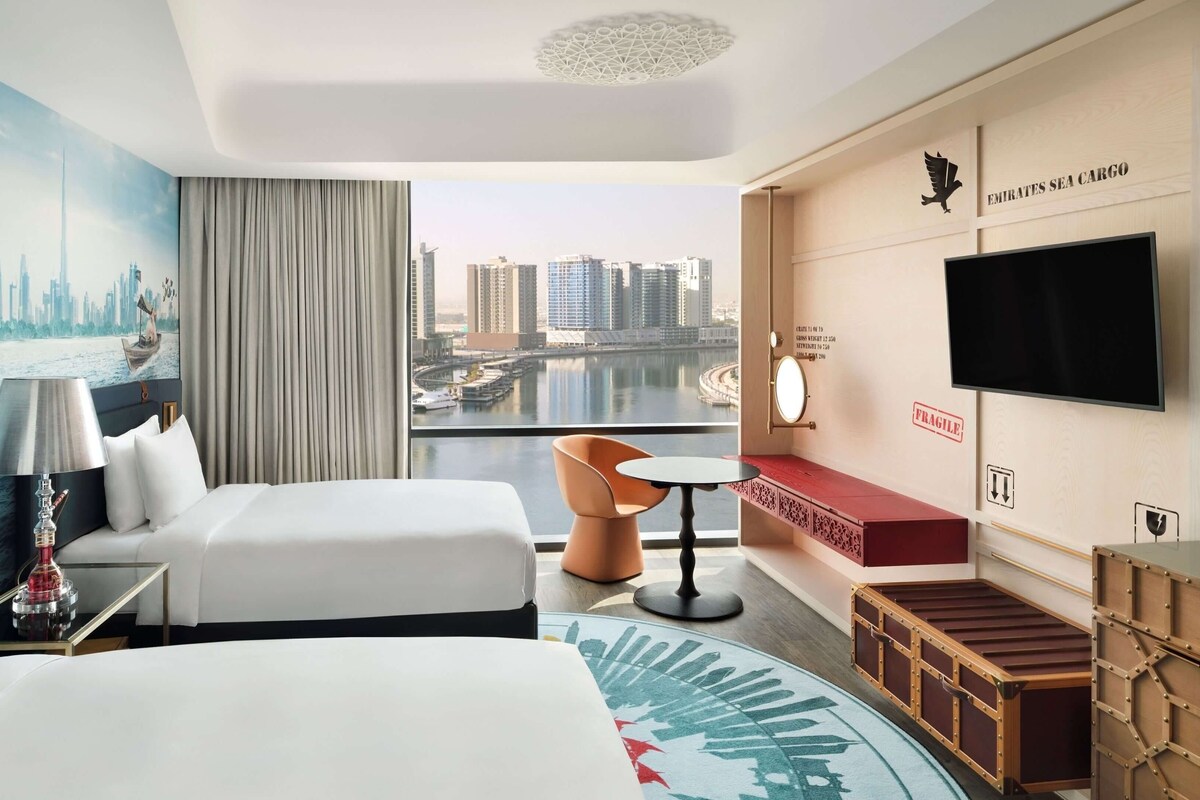 Ein Hotelzimmer mit einem Bett, zwei Nachttischen, einem Fernseher, einem Schreibtisch, einem Sessel und Blick auf die Stadtlandschaft.
