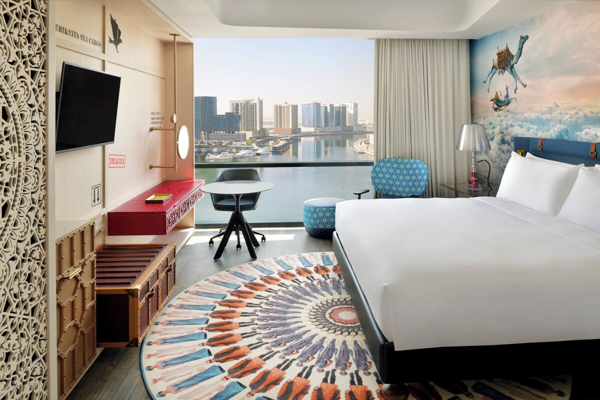 Ein Hotelzimmer mit einem großen Bett, einem Schreibtisch mit Stuhl, einem kleinen Tisch, einem an der Wand befestigten Fernseher und Blick auf die Stadt.
