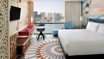 A hotel room with a large bed, a desk with a chair, a small table, a TV mounted on the wall, and a view of the cityscape through the window.