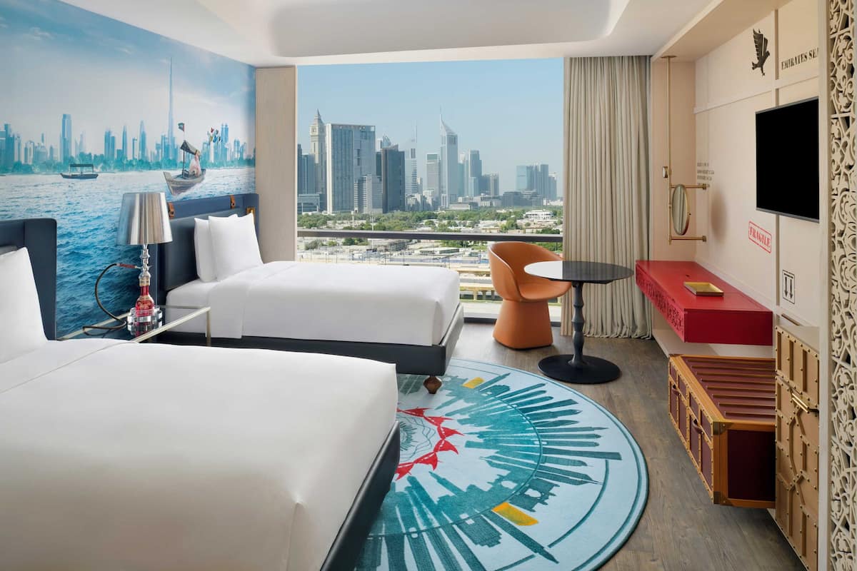 Ein Hotelzimmer mit zwei Betten, einem Stadtbild-Wandgemälde, Blick auf die Stadtlandschaft, einem kleinen Tisch und einem an der Wand befestigten Fernseher.