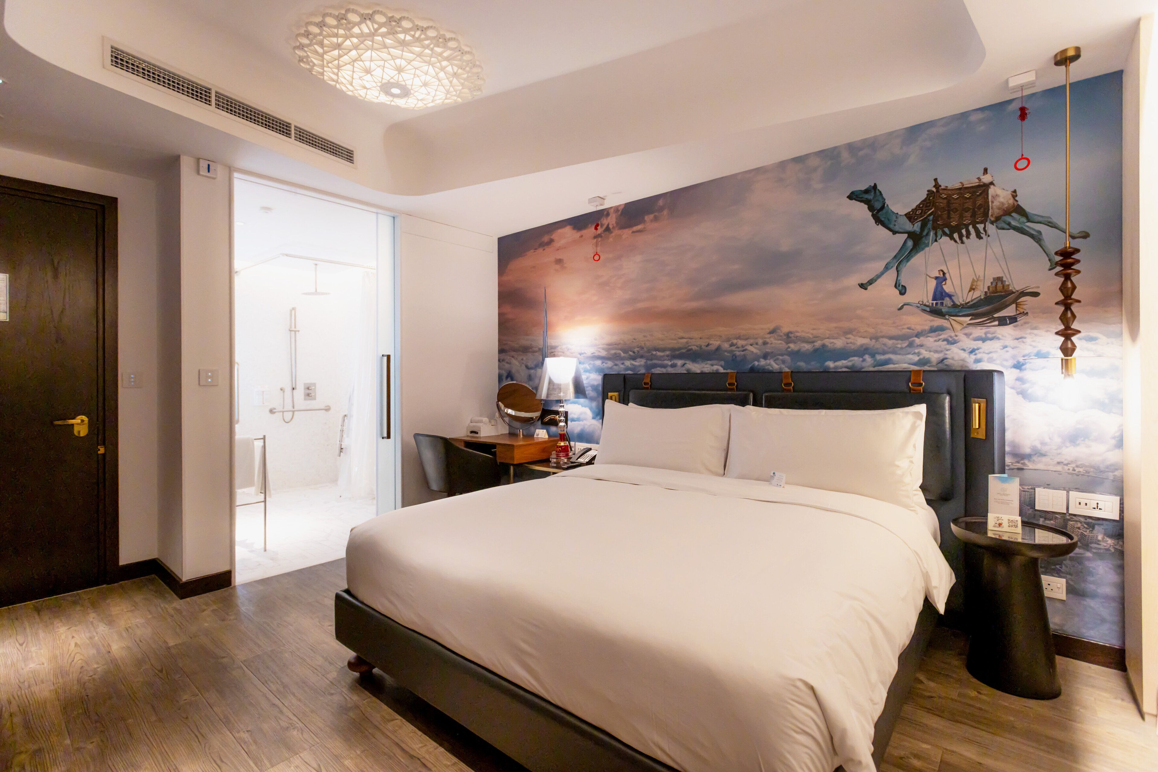 A hotel room with a large bed, a nightstand, a desk with a chair, and a wall mural of a camel on a boat.